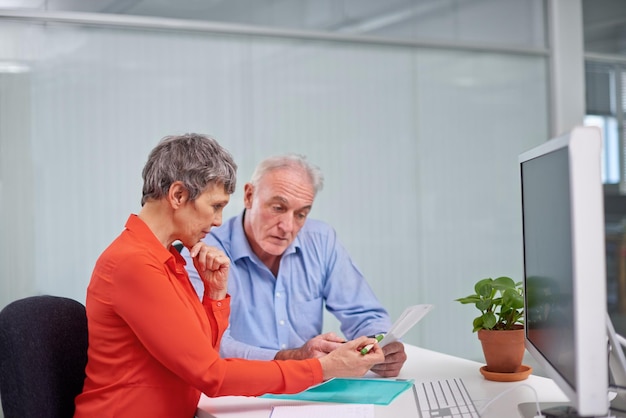 A government employee assisting an elderly person at a CRAS counter, demonstrating the process of applying for the Carteira do Idoso, with documents and a computer in view.