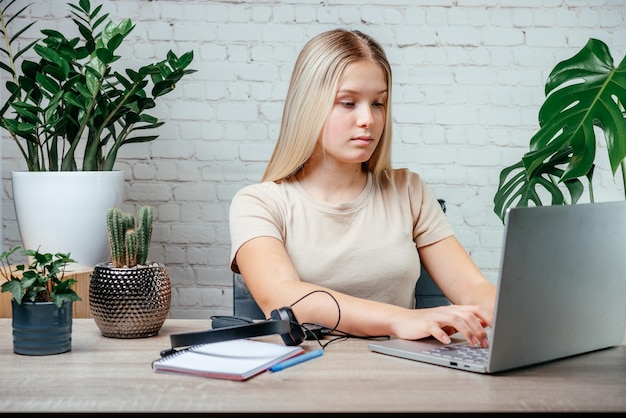 A young woman sitting comfortably at her desk, using a laptop to participate in an interactive online course. She is smiling and looks engaged, with a notebook and pen nearby. The background is a cozy, well-lit home office environment.